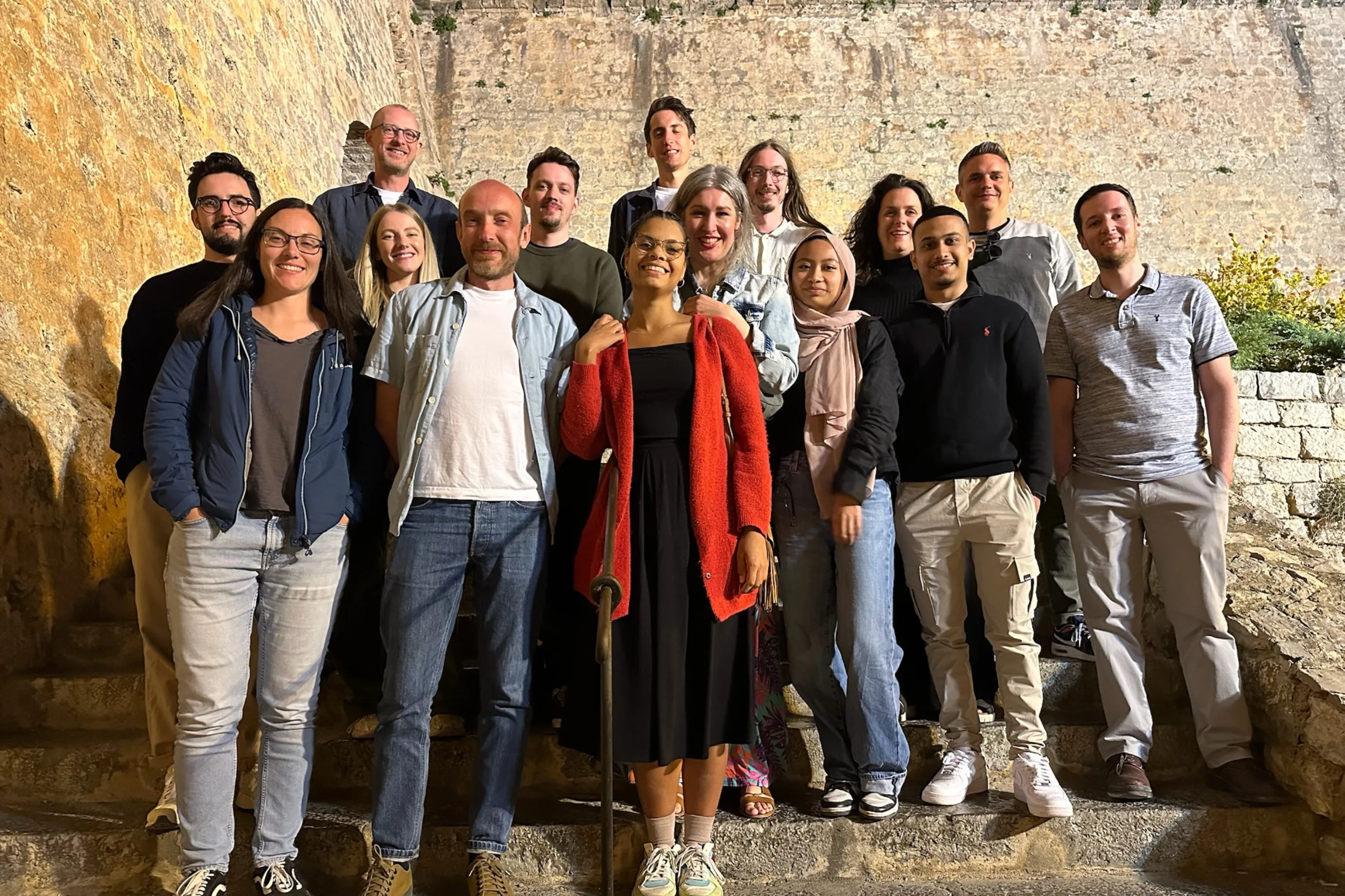 6 women and 9 men pose for a team photo on old stone stairs, with fortified stone walls in the background