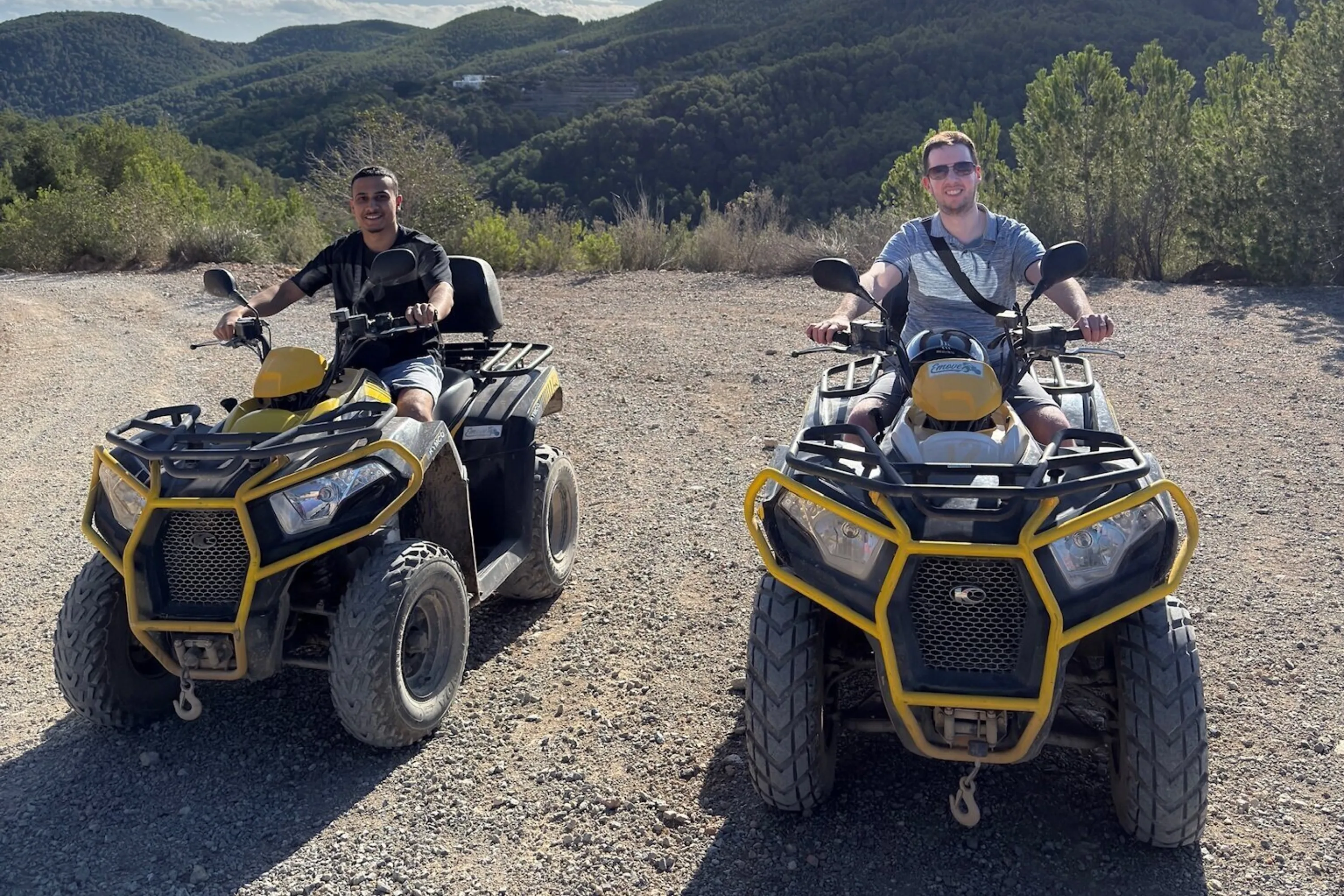 Two men pose for a photo on quad bikes with forested hills in the background