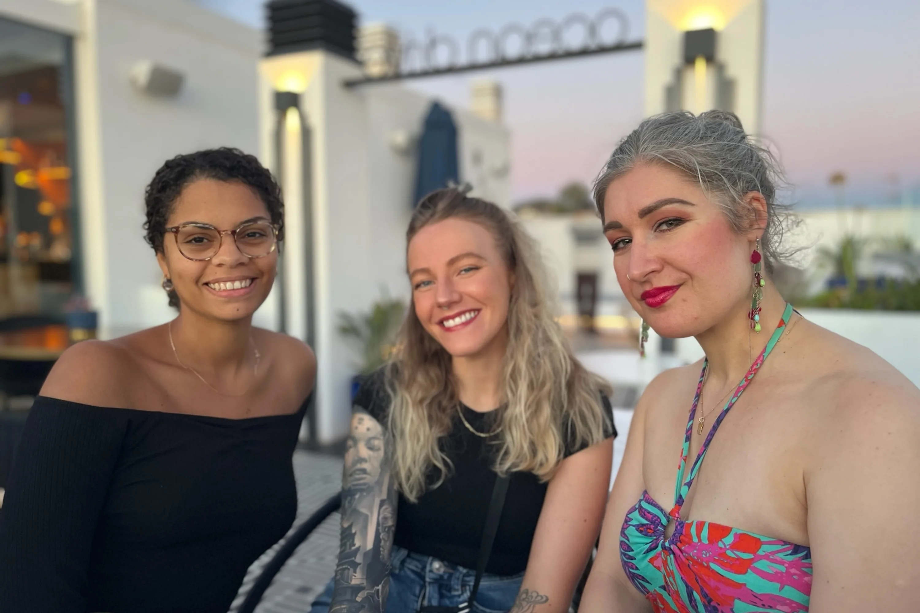 Three women smile and pose for a photo on a hotel rooftop bar at sunset
