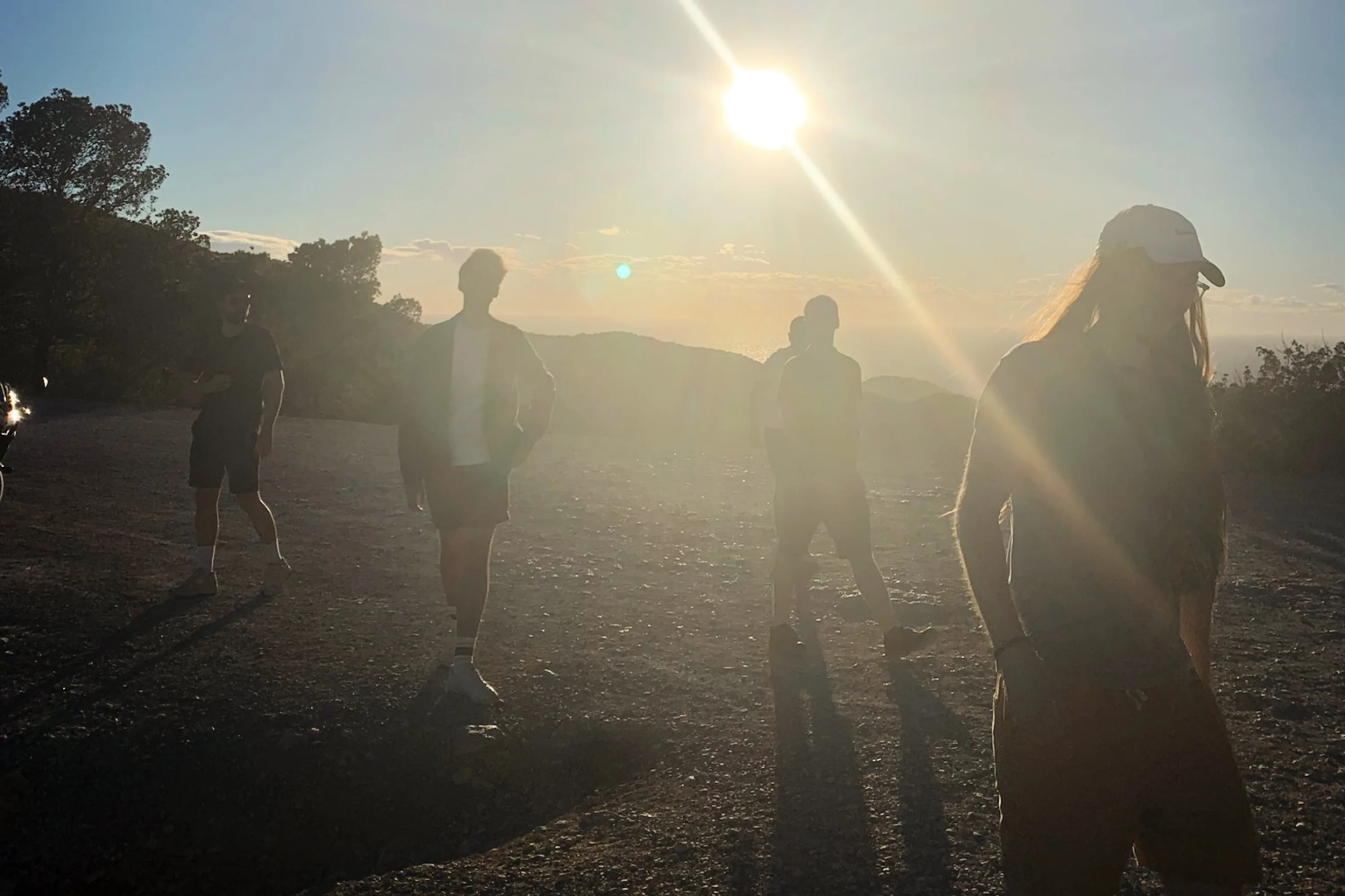 Five figures in semi-silhouette walk towards the camera as the sun sets behind them on a hilltop.