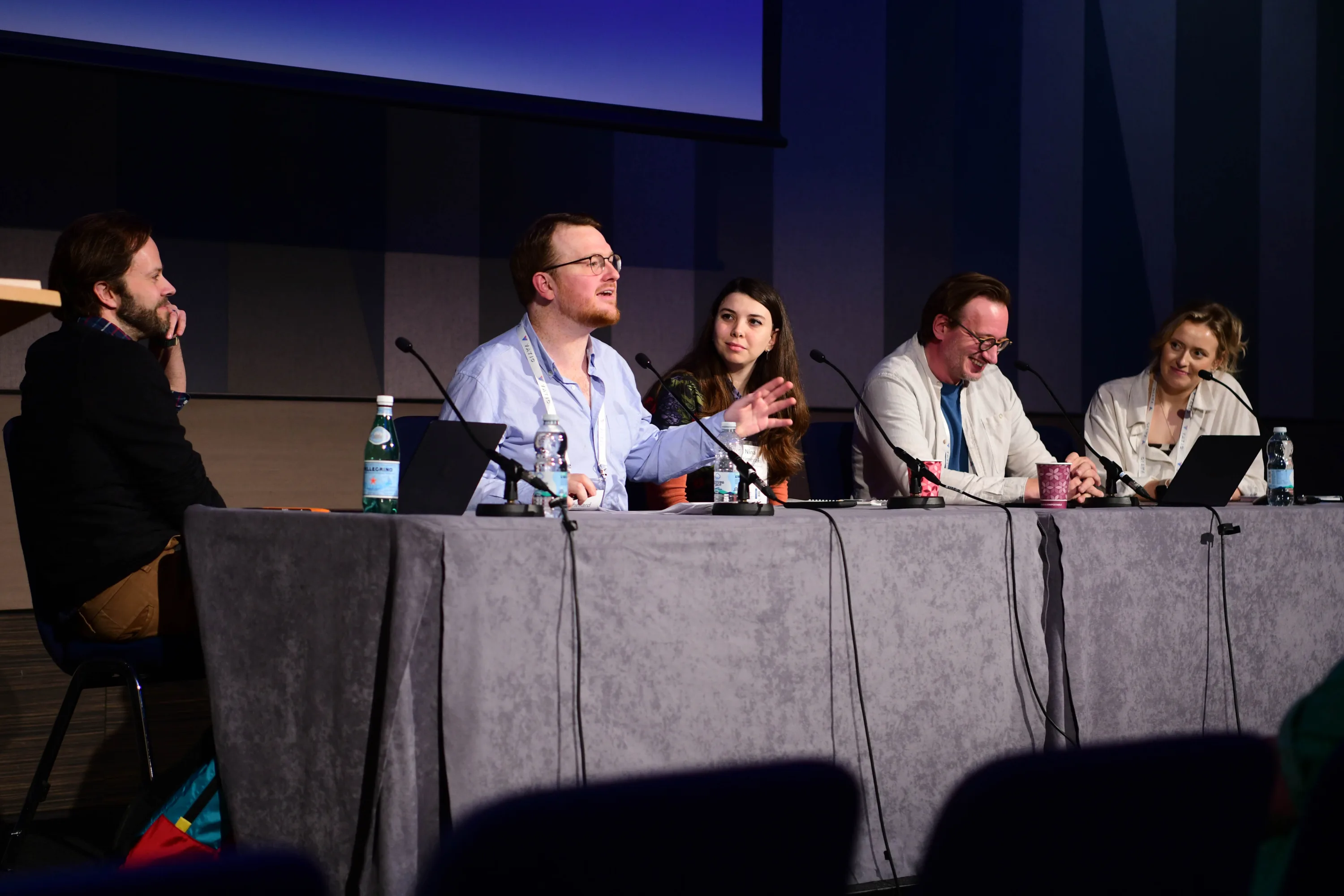 Five people sit on stage, each has a microphone. One is speaking while the others listen.
