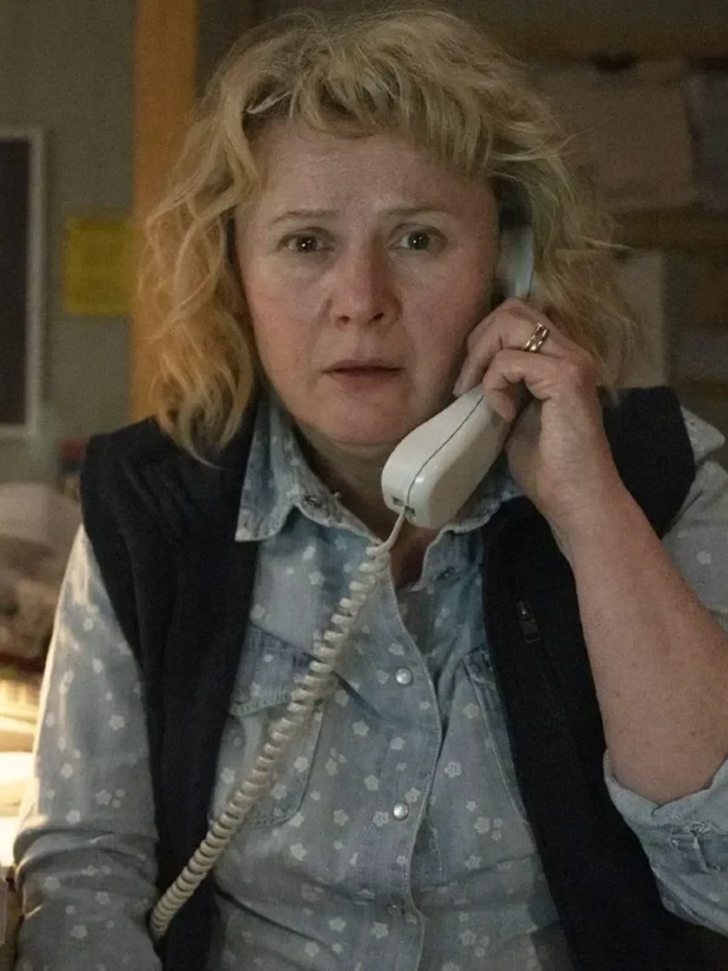 A middle aged woman looks worried holding a telephone to her ear in a small dimly lit office