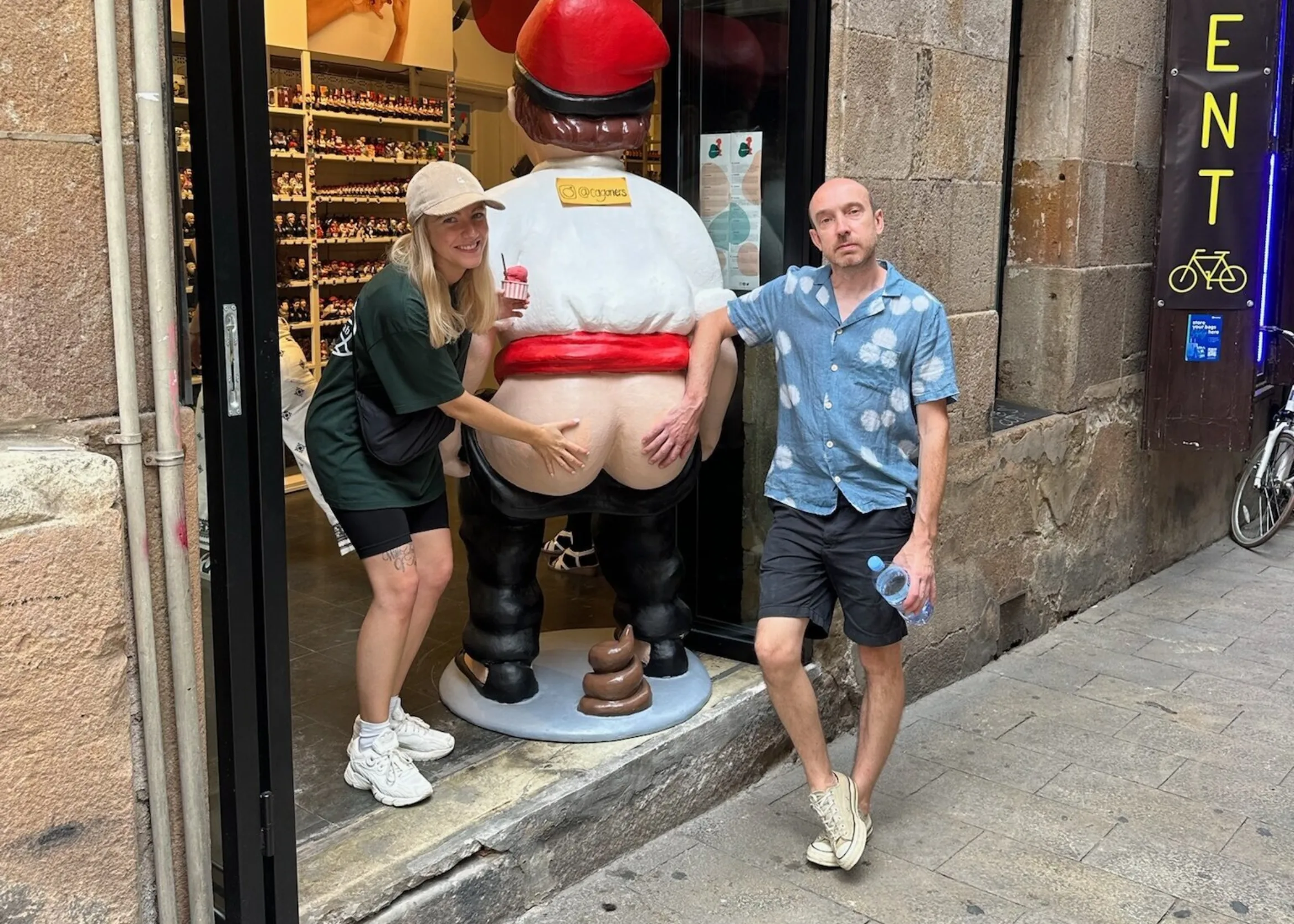 A man and a woman each holding a bare buttock of a large figurine, the figurine has defecated on the floor.