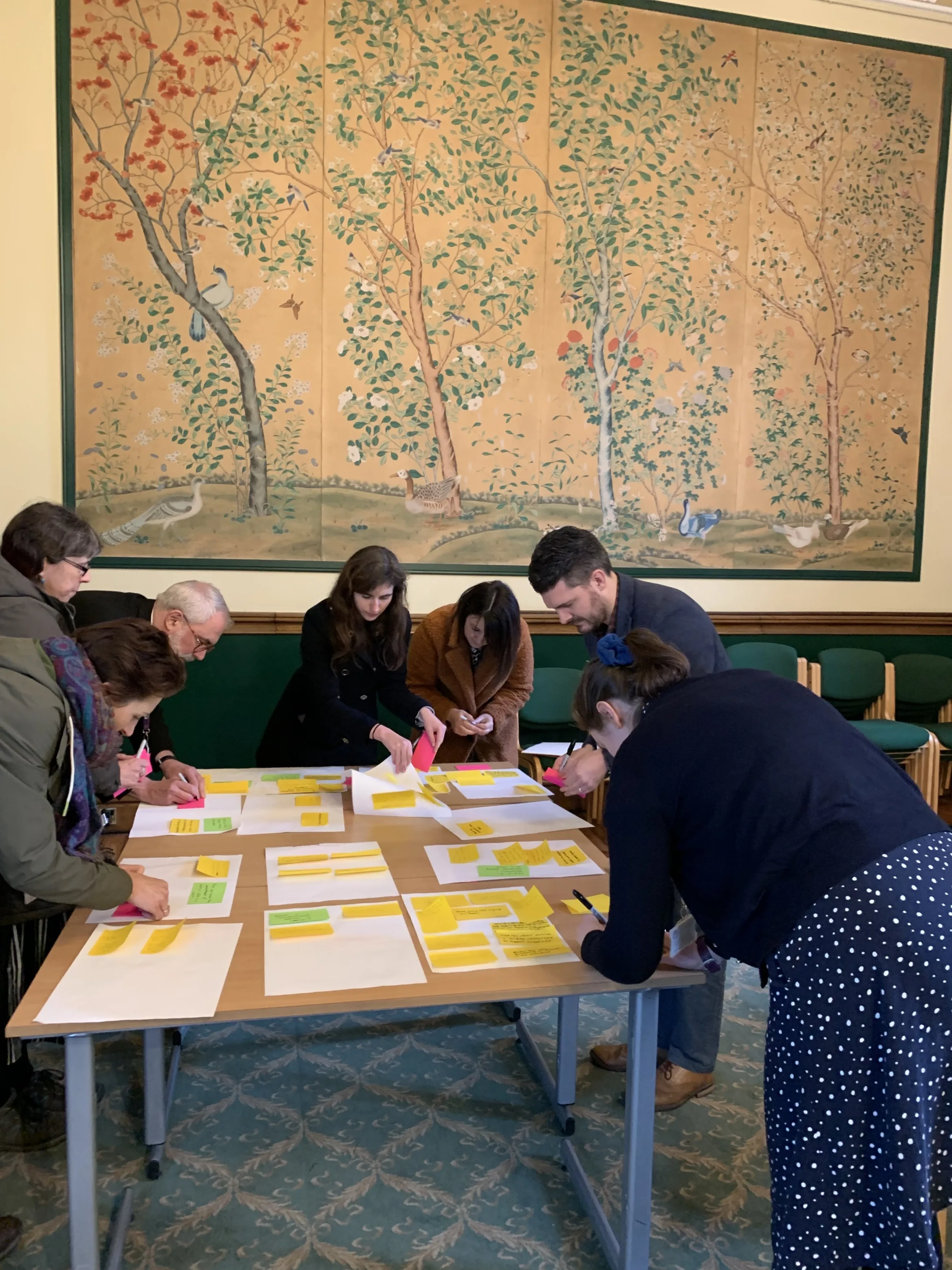 People standing around a central table placing post-it notes on larger pieces of paper with a large floral mural in the background