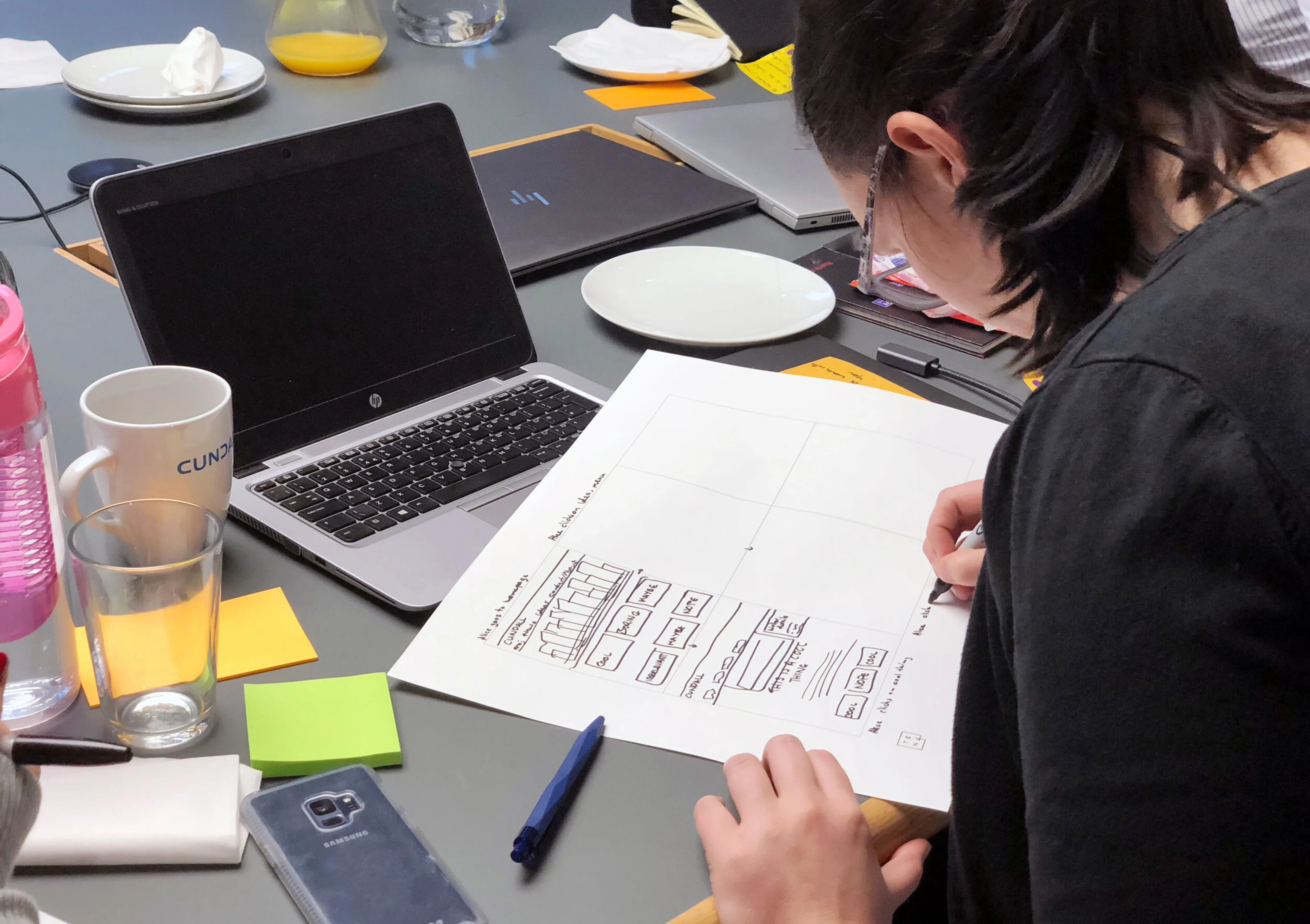 A woman sits at a busy desk and sketches ideas for website layouts