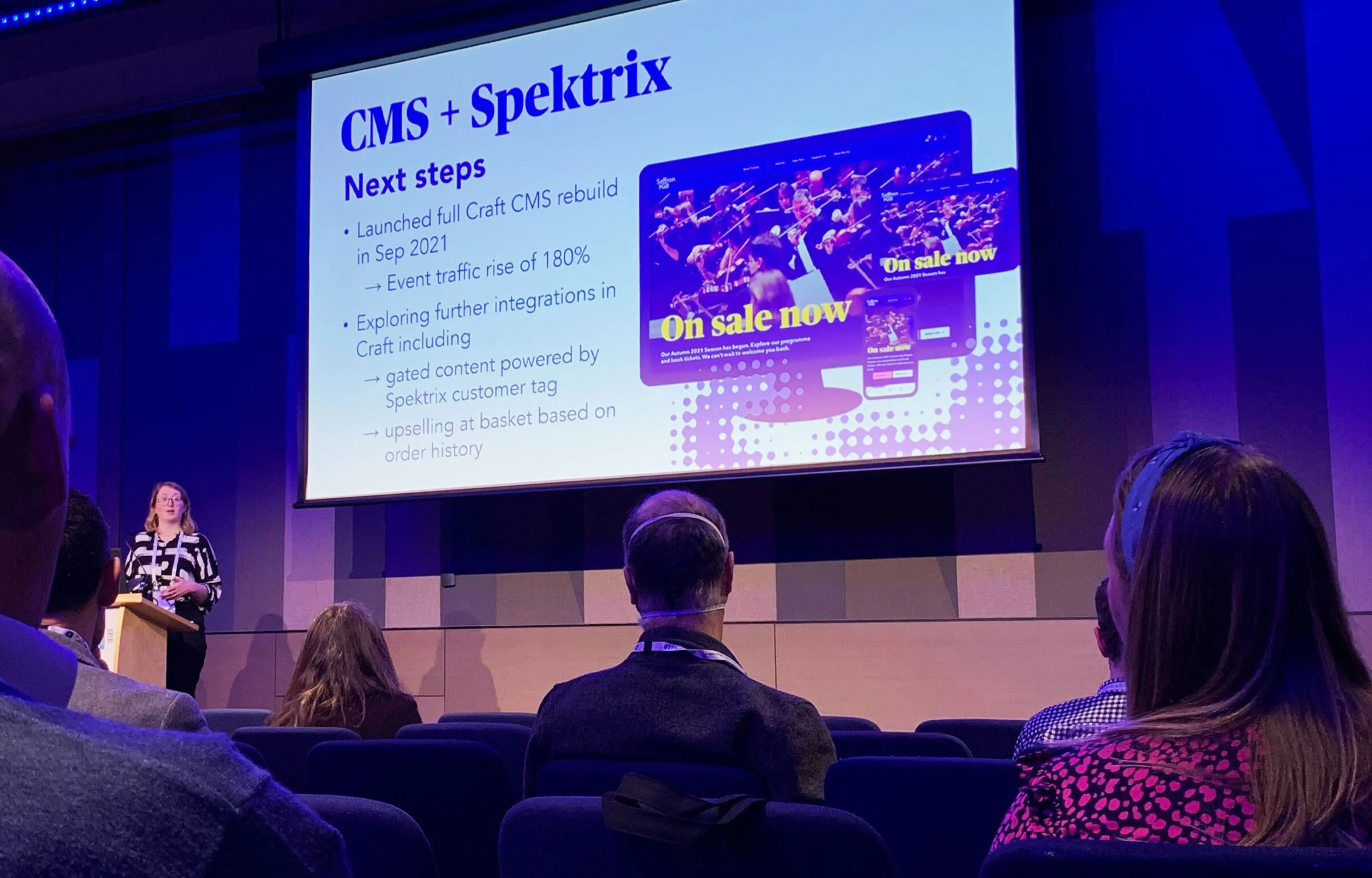 Emma Hall speaking on stage with a large screen behind. The screen is titled "CMS + Spektrix: Next steps" and shows screenshots from Saffron Hall's website.