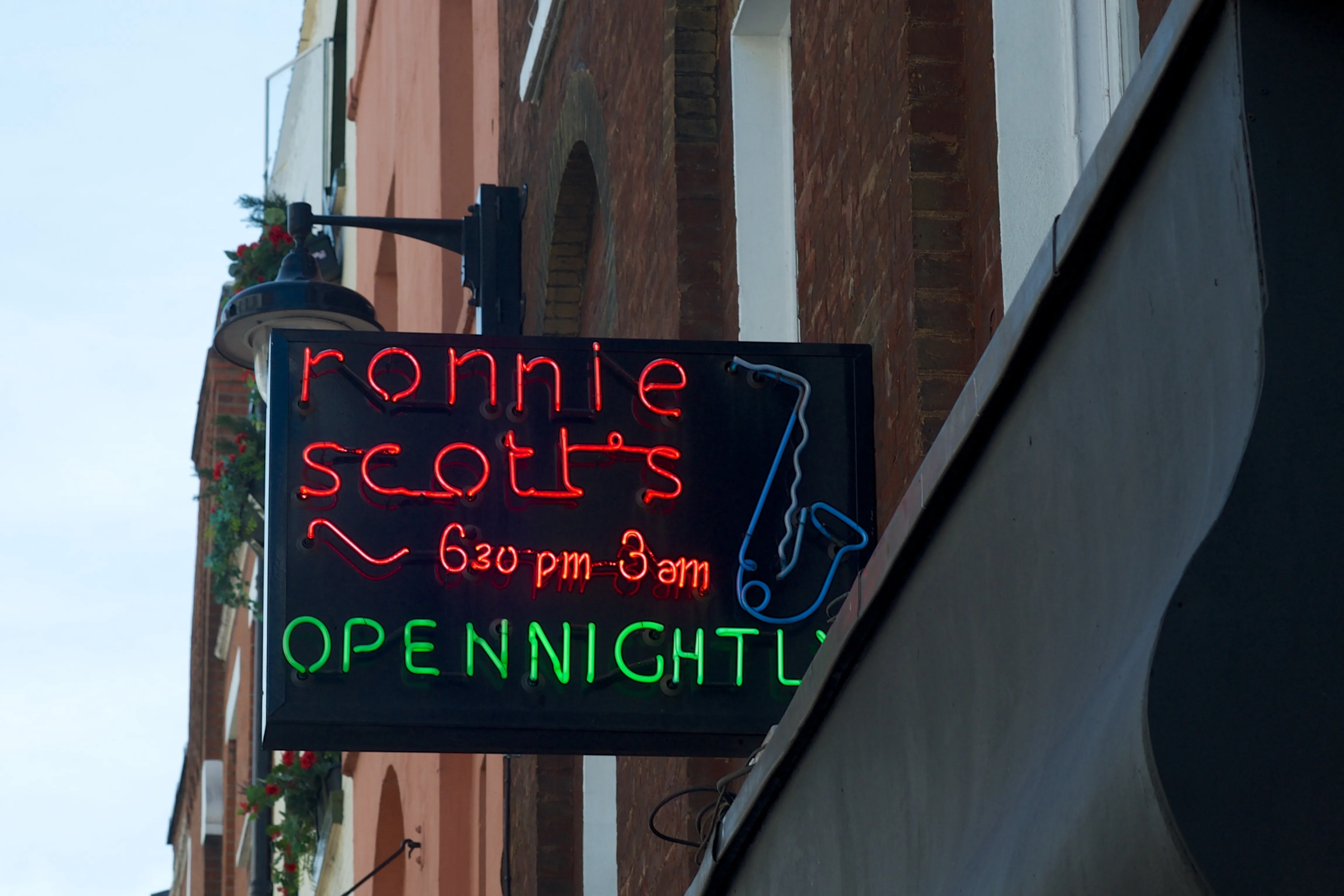 A neon sign with an outlines saxophone and the words "ronnie scott's 6:30pm–3am pen nightly"